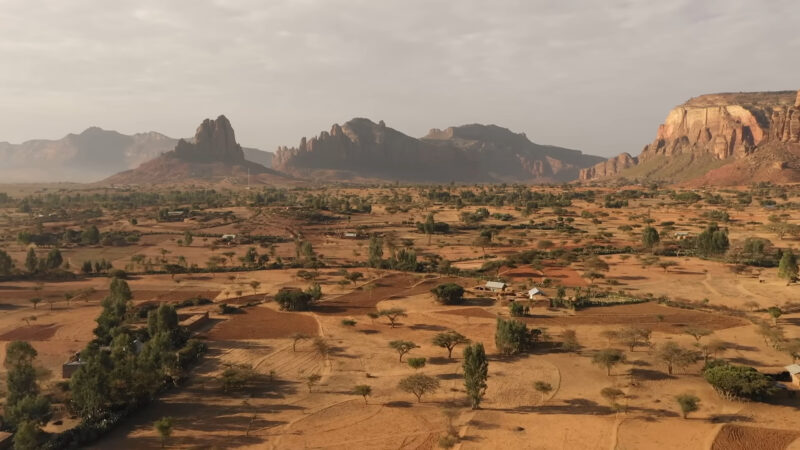 landscape view of Ethiopia, showcasing a vast, dry terrain dotted with small trees and traditional huts, with dramatic rocky mountains and cliffs in the background under a partly cloudy sky.