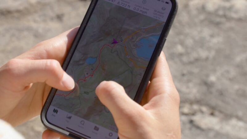 A close-up of a person's hands holding a smartphone displaying a GPS navigation app with a hiking trail map